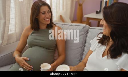 Schwangere Frau sitzt auf dem Sofa mit einer anderen Frau, die Kaffeetassen in einem gemütlichen Wohnraum hält Stockfoto