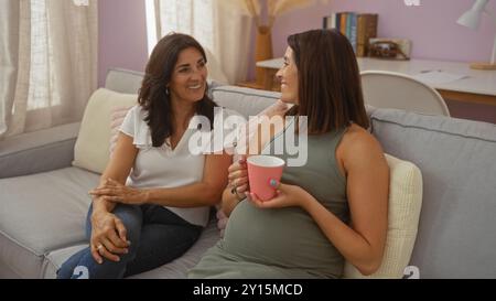 Schwangere Frau, die eine Kaffeetasse hält, während sie auf einer Couch sitzt und mit einer anderen Frau in einem gemütlichen Wohnzimmer spricht Stockfoto