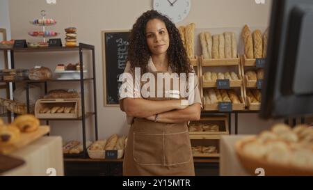 Die hispanische Frau mit lockigen Haaren und überkreuzten Armen steht selbstbewusst in einer Bäckerei mit verschiedenen Broten und Backwaren und präsentiert ein Indoor-Geschäft envi Stockfoto