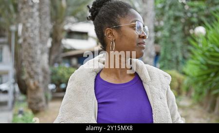 Eine kontemplative afroamerikanerin in einem Stadtpark mit Brille und einer gemütlichen Jacke. Stockfoto