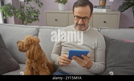 Ein Mann mittleren Alters entspannt sich mit seinem Pudel auf einer Couch zu Hause und schaut sich eine Tablette an Stockfoto