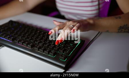 Nahaufnahme der Hand einer jungen Frau mit roten Nägeln, die auf einer hinterleuchteten Spieltastatur in einem dunklen Raum tippen Stockfoto