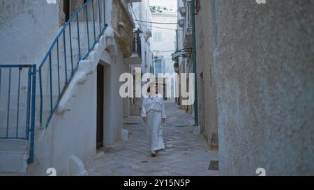 Eine junge, schöne hispanische Frau spaziert durch die charmanten gepflasterten Straßen von polignano a Stute, apulien, italien, in weiß gekleidet und umgeben von B Stockfoto