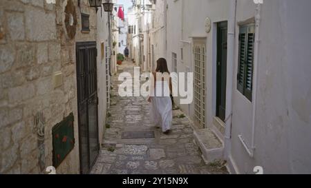 Eine junge, wunderschöne hispanische Frau in einem weißen Kleid schlendert durch die bezaubernden engen Gassen der ostuni Altstadt in apulien, italien, europa. Stockfoto