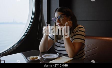 Besinnliche Frau, die Kaffee in einem Kreuzfahrtschiff-Café genießt und aus dem Bullauge auf dem Meer blickt. Stockfoto