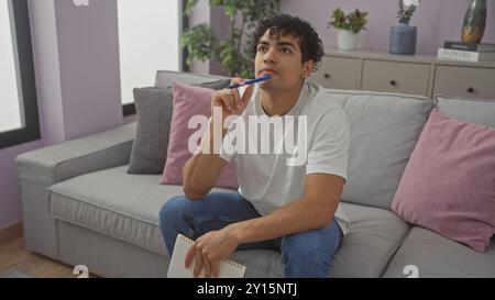 Ein nachdenklicher junger hispanischer Mann sitzt nachdenklich auf einer grauen Couch in einem modernen Wohnzimmer und hält einen Stift und ein Notizbuch. Stockfoto