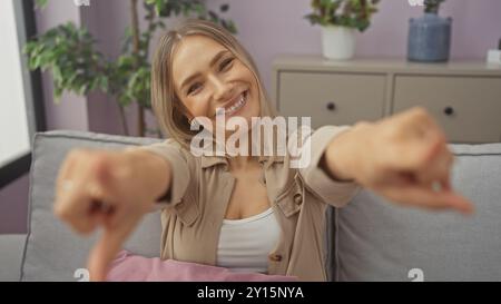 Eine schöne, blonde, junge Frau lächelt drinnen in einem gemütlichen Wohnzimmer und zeigt spielerisch mit Pflanzen im Hintergrund. Stockfoto