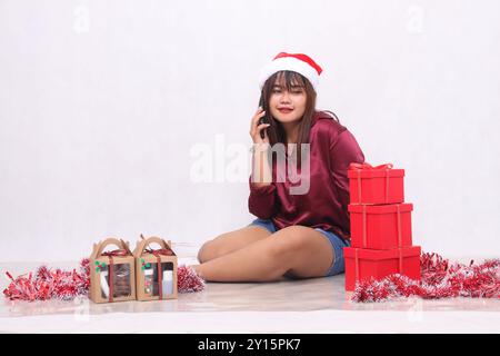 Schönes junges südostasiatisches Mädchen lächelt am Telefon neben 3 Geschenkschachteln an Weihnachten mit Weihnachtsschmuck und rotem Hemd auf weißem Hintergrund Stockfoto