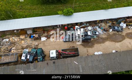Ein Blick aus der Vogelperspektive auf einen Schrottplatz mit verschiedenen demontierten Autos und Teilen, die auf einem unbefestigten Weg verstreut sind. Die Gegend ist von Grün umgeben und hat einen Stockfoto