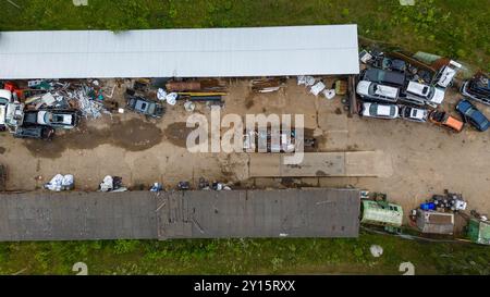 Ein Blick aus der Vogelperspektive auf einen Schrottplatz mit verschiedenen demontierten Autos und Teilen, die auf einem unbefestigten Weg verstreut sind. Die Gegend ist von Grün umgeben und hat einen Stockfoto