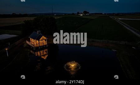 Ruhige Farm-Szene in der Abenddämmerung mit Einem gemütlichen Steinhaus, beleuchtet von sanfter Beleuchtung, reflektiert in Einem friedlichen Teich mit Einem Brunnen, umgeben von weitläufigen grünen Feldern Stockfoto