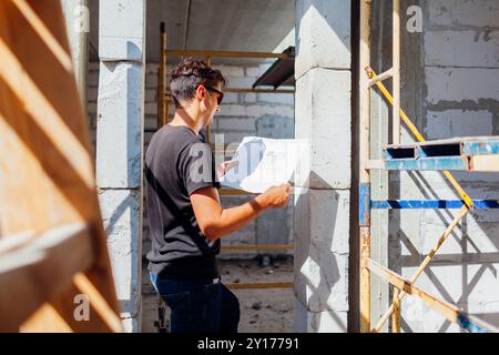Mann Architekt Ingenieur überprüft Haus im Bau und vergleicht mit dem Plan auf Papier, das an Leiter und Gerüst steht. Projektgebäude mit Stockfoto