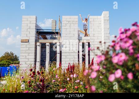 Glückliche junge Frau, die ihr Haus im Bau überprüft, das aus Gasblock besteht und im ersten Stock neben dem Fensterschlitz steht. Der zukünftige Besitzer stellt sich ein gemütliches Zuhause vor Stockfoto