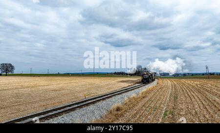 Eine historische Dampfeisenbahn schlängelt sich entlang der Gleise und weht weißen Rauch, während sie durch offene Felder unter einem dramatischen Himmel voller Wolken schneidet. Stockfoto