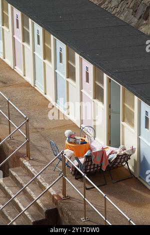 Eine Person, die sich auf Klappstühlen neben Einem Tisch vor Einer Strandhütte am Sidmouth Beach, Sidmouth, Großbritannien, entspannt Stockfoto