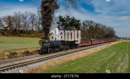 Eine alte Dampflokomotive spritzt schwarzen Rauch ab, während sie entlang der Eisenbahngleise schlummert, umgeben von üppigen grünen Feldern und Bäumen unter einem klaren blauen Himmel. Passagiere genießen die malerische Fahrt. Stockfoto