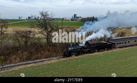 Eine alte Dampflokomotive zieht einen Personenzug entlang einer landschaftlich reizvollen Strecke, umgeben von Feldern und entfernten Scheunen. Der Himmel ist bedeckt und verleiht der malerischen Landschaft ein stimmungsvolles Ambiente. Stockfoto