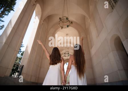 Zwei Frauen in weißen Kleidern stehen in einem bogenförmigen Eingang, eine mit erhobenem Arm. Sie blicken von der Kamera weg und blicken auf einen Kronleuchter. Stockfoto