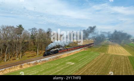 Eine Dampflokomotive zieht einen alten Zug durch eine malerische Landschaft und strahlt Rauch und Dampf vor der Kulisse von Feldern und Bäumen unter hellem Himmel aus. Stockfoto