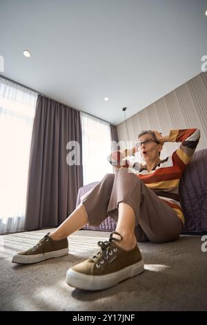 Eine verängstigte Seniorin in Brille sitzt auf dem Boden neben dem Bett im Hotel Stockfoto
