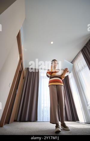 Die ganze Länge einer älteren Frau, die mit einem Laptop arbeitet und im Hotelzimmer über ein Smartphone spricht Stockfoto