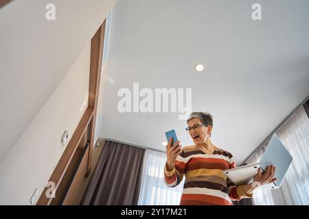 Ältere Frau, die auf das Smartphone schaut, während sie schreiend im Hotel ihren Laptop hält Stockfoto