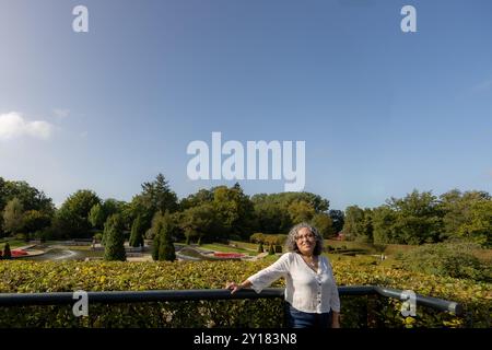 Erwachsene Frau, die auf der Aussichtsterrasse mit Innengärten von Arcen Castle im Hintergrund steht, Brunnen und üppige Bäume, lässige Kleidung, weiße Bluse, sunn Stockfoto