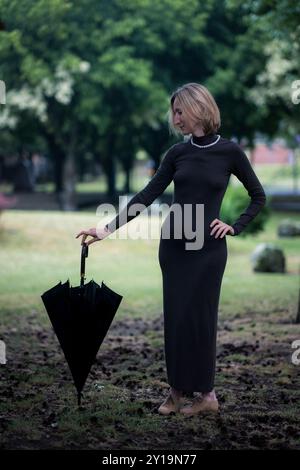 Eine Frau mit einem zusammengefalteten Schirm steht an einem bewölkten Tag in einem Stadtpark. Stockfoto