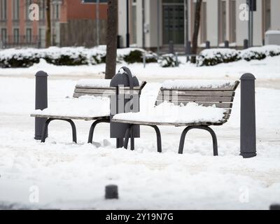 Leere Bank mit Schnee drauf. Sitzplatz in der Fußgängerzone neben dem Bürgersteig während der Wintersaison. Weißer Schnee bedeckt die städtische Umgebung. Stockfoto