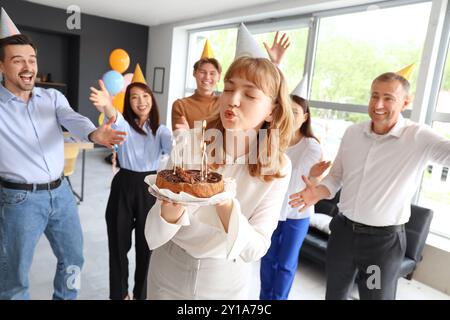 Junge Geschäftsfrau, die Kerzen auf Kuchen auf der Geburtstagsfeier im Büro ausbläst Stockfoto