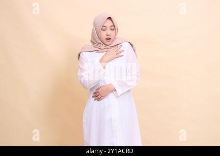 Eine asiatische Hijab-Frau, die ein weißes Kleid und einen braunen Schal trägt, steht und schließt ihre Augen mit den Händen auf der Brust. Für Gesundheit, Pflege, educ Stockfoto