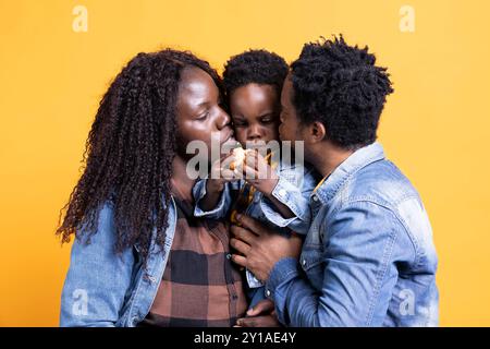 Liebe afroamerikanische Eltern, die ihren kleinen Sohn vor der Kamera küssen und Glück mit ihrer kleinen Familie haben. Ein junges Paar umarmt ihren kleinen Jungen vor gelbem Hintergrund. Stockfoto