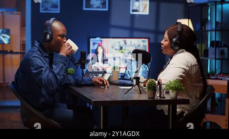 Zeigen Sie den Host, der Podcast aufnimmt, genießen Sie eine Tasse Kaffee und nette Gespräche mit den Gästen. afroamerikanischer Mann, der professionelle Audiogeräte für eine einwandfreie Klangqualität verwendet, während er mit einer Frau im Live-Chat spricht Stockfoto