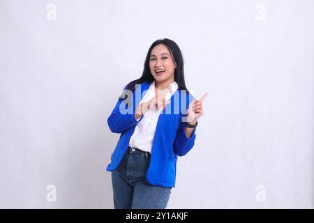 Der Charme eines hübschen jungen asiatischen Büromädchens in einem blauen formellen Anzug, der fröhlich steht und beide Hände nach oben links zeigen. Für corporat Stockfoto