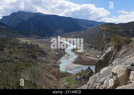 Ein Fluss schlängelt sich durch ein weites Tal, umgeben von grünen Hügeln und felsigen Bergen unter einem teilweise bewölkten Himmel, Noguera Ribagorcana Mont-rebei Natura Stockfoto