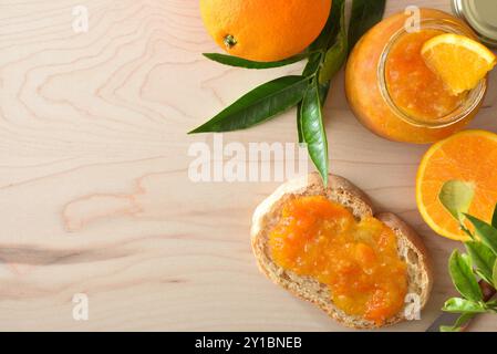 Hausgemachte Orangenmarmelade im Glasgefäß und Toast auf Holztisch mit Obststücken herum. Draufsicht. Stockfoto