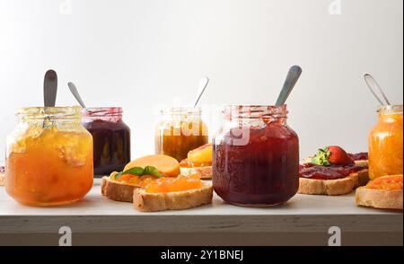 Gläser voller Marmelade mit verschiedenen Geschmacksrichtungen und Brotscheiben mit Marmelade dekoriert und isoliertem weißen Hintergrund. Vorderansicht. Stockfoto