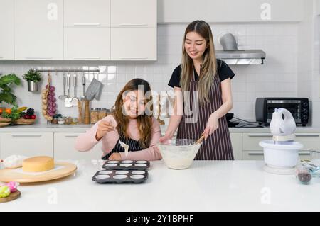 Zwei junge Frauen machen Nachtisch in der Küche. Eine Person, die einen weißen Kuchenteig oder eine Glasmischung in einer klaren Glasschale umrührt. Der andere Teig wird gegossen Stockfoto