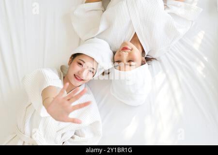 Zwei asiatische Frauen liegen in weißen Bademänteln und Handtüchern um den Kopf gewickelt auf einem Bett. Draufsicht Stockfoto