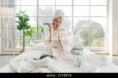 Junge asiatische Frau sitzt entspannt auf einem Bett in weißem Bademantel und Handtuch um ihren Kopf gewickelt, schminkt sich mit einem Spiegel und einem Schminkkkoffer mit Reißverschluss. Stockfoto