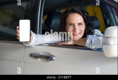 Die weiße Frau sitzt mit dem Fenster nach unten auf dem Fahrersitz eines Autos. Sie lächelt und hält ein Smartphone mit einem leeren Bildschirm in Richtung C Stockfoto