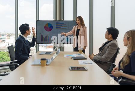 Ein junger asiatischer Geschäftsmann in Anzug hebt die Hand, um eine Frage an den Moderator zu stellen, der vor einem großen digitalen Monitor steht. Führungskräfte Team m Stockfoto