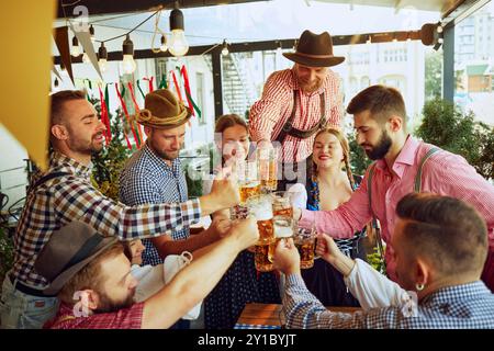 Applaus zur deutschen Tradition, Gruppe von Menschen in bayerischen und legeren Klamotten, Freunde treffen sich im Pub, Bierbecher in feierlichen Toasts Stockfoto