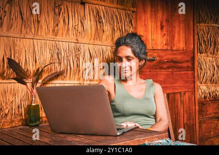 Hipster-Frau fokussierte Arbeit online in abgelegenen tropischen Dorf isoliert sitzen am Bungalow mit Computer umgeben von grüner üppiger asiatischer Natur Stockfoto