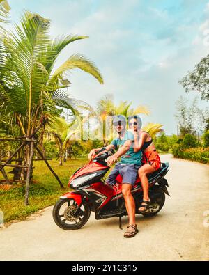 Glückliches lächelndes Paar Reisende, die während eines tropischen Urlaubs unter Palmen auf der Insel Phu Quoc, Vietnam, mit einem Motorroller in einem Sicherheitshelm fahren Stockfoto