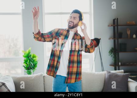 Foto von hübschen jungen Mann Tanz Kopfhörer hören Musik gekleidetes kariertes Hemd Verbringen Sie Zeitvertreib geräumiges Haus im Zimmer Stockfoto