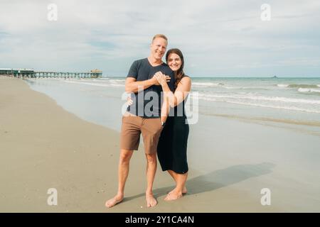 Ein Paar steht an einem Sandstrand in der Nähe eines Piers, umschmeichelt und lächelt Stockfoto