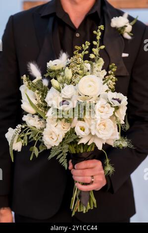 Bräutigam in schwarzem Smoking, der den weißen Hochzeitsstrauß der Braut hält Stockfoto