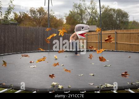 Junge im Haifischkostüm auf Trampolin mit Herbstlaub an bewölktem Tag Stockfoto