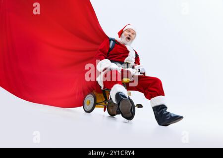 Weihnachtsmann mit weißem Bart reitend Dreirad mit riesigen roten Geschenktüten hinter sich, die vor weißem Studiohintergrund fließen. Stockfoto